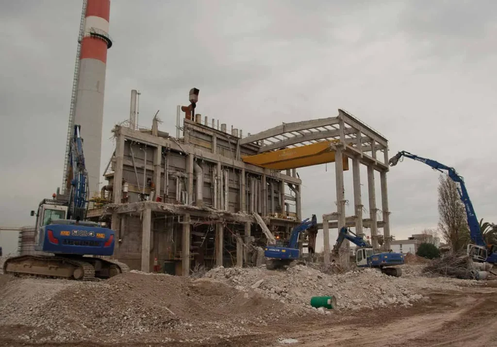 Retroexcavadoras de demolición derribando una estructura industrial de hormigón junto a una chimenea